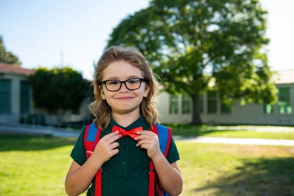 Kind mit Rucksack im Schulpark. Schüler mit Rucksäcken im Freien. Nerd-Schüler bindet Fliege, korrigiert rote Fliege. — Stockfoto