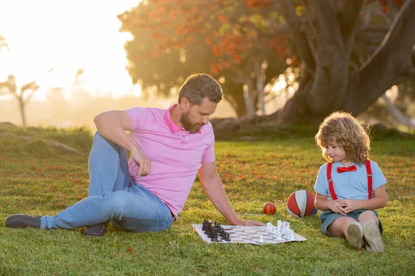Kid son sitting on grass and playing chess with father. Clever thinking smart child while playing chess.