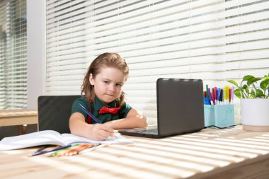 Sevimli okul çocuğu ödevini evde tablet ya da dizüstü bilgisayarla yapıyor. Öğrenci görevi bir deftere yazar. Eğitim ve öğrenim, çocuklar evde eğitim..