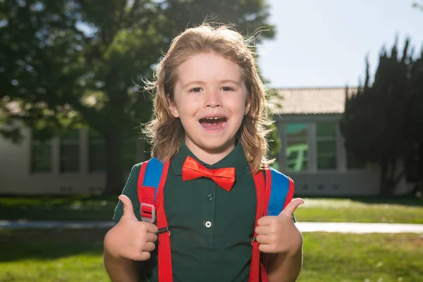 Alegre niño de escuela en uniforme escolar con gran mochila de pie cerca de la escuela. De vuelta a la escuela. Concepto de educación preescolar infantil. —  Fotos de Stock