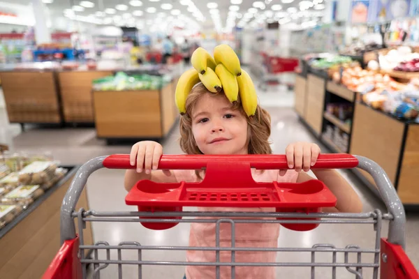 Banana on funny child head. Funny kid with shopping cart buying food at grocery store or supermarket. Surprised kids face. — Stock Photo, Image