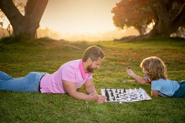 Figlio sdraiato sull'erba e giocare a scacchi con il padre al tramonto nel parco soleggiato. — Foto Stock