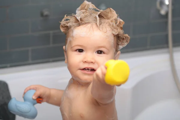 Ευτυχισμένο μωρό που κάνει μπάνιο παίζοντας με φυσαλίδες αφρού. Ένα μικρό παιδί στην μπανιέρα. Χαμογελώντας παιδί στο μπάνιο με πάπια παιχνίδι. Βρεφικό πλύσιμο και μπάνιο. Φροντίδα και υγιεινή των παιδιών. — Φωτογραφία Αρχείου