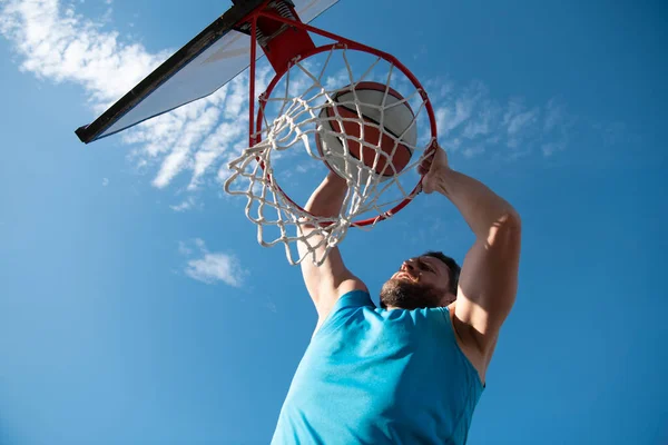 Basketbalspeler. Sport en basketbal. Een jongeman springt en gooit een bal in de mand. Blauwe lucht en hof op de achtergrond. — Stockfoto