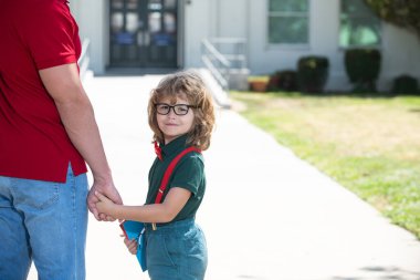 Öğretmenlerin elini tutan mutlu inek öğrenci portresi. Okullu çocuk babamla aynı okula gidiyor. Okul kapısının önünde çocuklu bir ebeveyn.