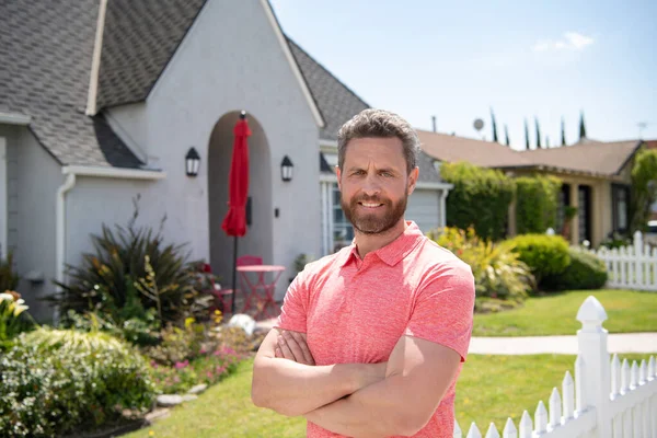 Um americano sorridente com braços cruzados em frente à sua nova casa. Compra, venda, imobiliário, propriedade, seguro de casa conceito. Pessoas reais. — Fotografia de Stock