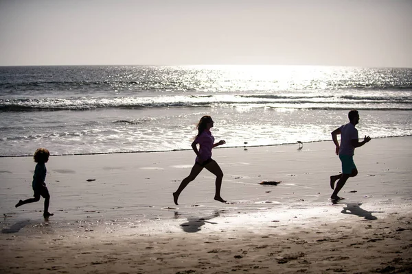 お父さん、お母さん、お子さんが海岸を走っています。健康的な家庭生活をコンセプトとする。夏の人が走り、親と子供が海でスポーティな休暇を過ごす。子供と一緒に屋外でジョギングする家族. — ストック写真