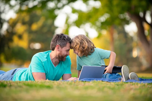 Boldog apa fiával használja a laptopot. Apa és iskolás fiú nézik a képernyőt és táblagépet, videóórát néznek, füvön ülnek. Családi hétvége. Az unoka videohívása nagyszülőknek. — Stock Fotó