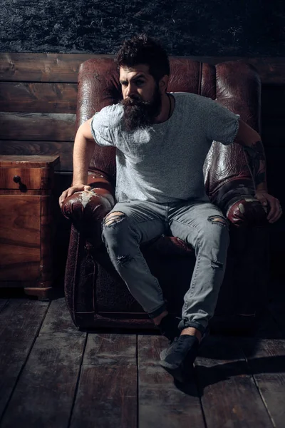 Handsome confident bearded man sitting in luxury leather chair and looks grumpy. Stylish attractive man with dramatic face. Bearded man wearing informal clothes and sitting in chair. — Stock Photo, Image