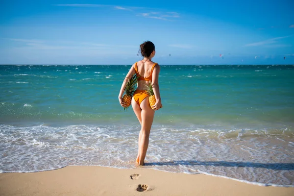 Femme en maillot de bain avec les fesses musculaires tenir l'ananas près de la plage de sable de l'île. Fruits sexy d'été. Joyeuse période chaude estivale de jeunes femmes heureuses avec de l'ananas. Modèles sexy en vacances tropicales. — Photo