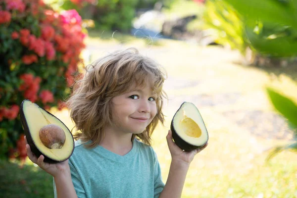 Närbild ansikte unge med en halv avokado i hennes handat tropisk trädgård. Friska barn avokado mat koncept. — Stockfoto