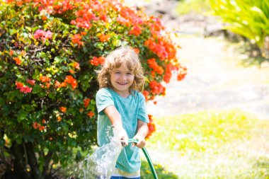 Yaz günü bahçede bitkileri sulayan tatlı çocuk. Bahçede bahçe aletleri ve sulama hortumu olan bir çocuk. Çocuk bahçede eğleniyor. Çocuk şefkati ve çocukluk kavramı.