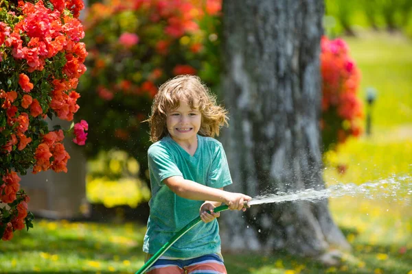 Child boy watering the plants, from hose spray with water hose in the garden at the backyard of the house on a summer evening. — Stock Photo, Image