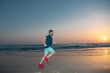 Spor giyim sektöründe erkek model plajda açık havada egzersiz yapıyor. Koşan atletik genç adam, günbatımında ya da gündoğumunda spor modeli.