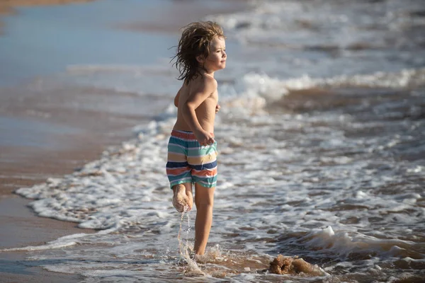 Niño corriendo en la orilla de la playa salpicando agua en el mar azul. Niño caminando por la playa de verano. — Foto de Stock