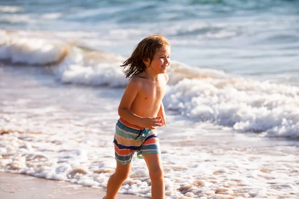 青い海に海水が飛び散る海岸を走っている少年。夏のビーチを歩く子供. — ストック写真