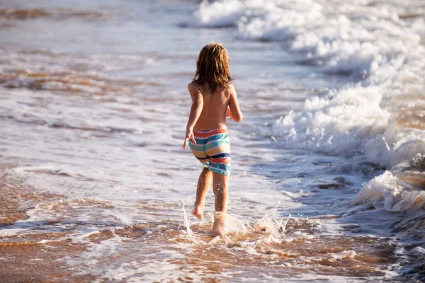 Vista trasera de un chico corriendo por la playa cerca del mar. Emocionado niño sorprendido divirtiéndose con correr a través del agua en el océano o el mar. — Foto de Stock