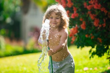 Amerikalı çocuk bahçede bahçe hortumuyla oynuyor. Komik heyecanlı çocuk arka planda su püskürterek eğleniyor. Yaz çocukları açık hava aktivitesi.