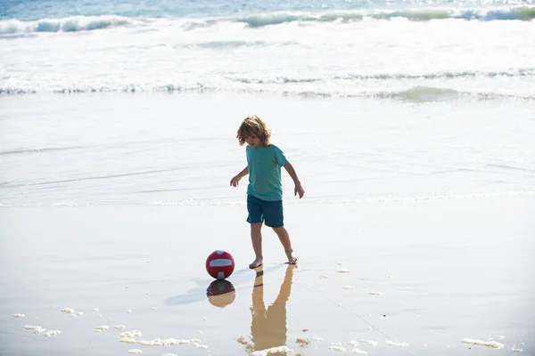 El niño juega al fútbol en la playa de arena. Fútbol de verano en la playa para niños. —  Fotos de Stock