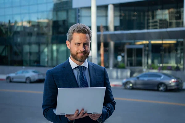 Geschäftsmann mit Notebook im Freien. Selbstbewusster Wirtschaftsexperte. Schöner Mann im Anzug hält Laptop vor Bürohintergrund. — Stockfoto
