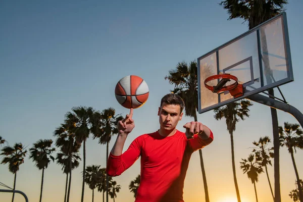 Ο αθλητής μπασκετμπολίστας με πουκάμισο και σορτς κρατάει μπάλα μπάσκετ σε αστικό φόντο. Αθλητικά κίνητρα. Χέρι περιστρεφόμενο καλάθι μπάλα. Εξισορρόπηση μπάσκετ στο δάχτυλο. — Φωτογραφία Αρχείου