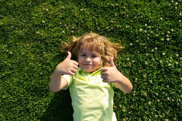 Criança engraçada com polegares para cima sinal deitado na grama, close up face. Crianças curtindo verão. — Fotografia de Stock