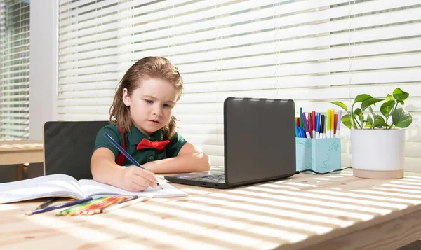 Schattig schooljongetje doet haar huiswerk met een tablet of laptop thuis. Pupil schrijft de taak in een notitieboekje. Onderwijs en leren, thuisonderwijs voor kinderen. — Stockfoto