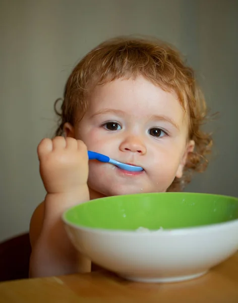 Ritratto di bambino caucasico carino con cucchiaio. Bambino affamato disordinato con piatto dopo aver mangiato purea. — Foto Stock