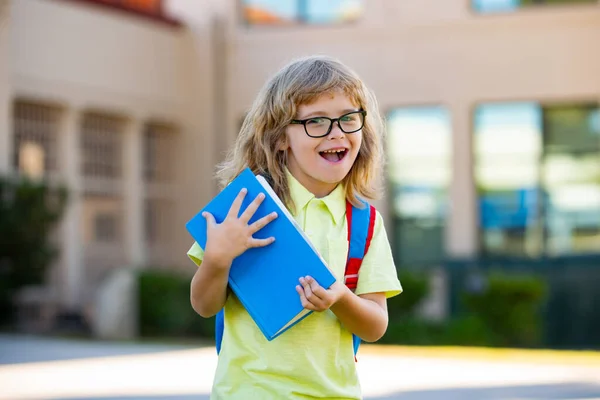 Büyük sırt çantalı neşeli küçük çocuk okula karşı eğleniyor. Okul konseptine dönelim.. — Stok fotoğraf