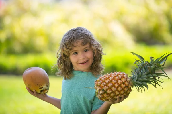 Retrato de un niño pequeño al aire libre en verano. Sonriente lindo chico divertido sosteniendo una piña y coco. — Foto de Stock