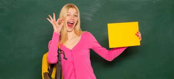 Spännande förbluffad lärare. Studerande flicka hålla tom blöja för produkt. Porträtt av leende kvinnlig lärare nära svarta tavlan i klassrummet i skolan. — Stockfoto