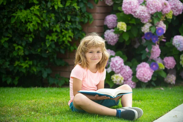 Junge liest ein Buch, das auf Gras liegt. Nettes kleines Kind in lässiger Kleidung, das ein Buch liest und lächelt, während es im Park auf Gras liegt. Lernkonzept. — Stockfoto