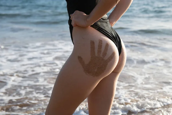 Ragazza con bottino sexy con stampe a mano in bikini nero appoggiato sulla spiaggia deserta. Bellissimo modello in costume da bagno sulla sabbia tropicale vicino al mare. — Foto Stock