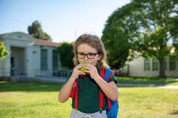Веселий маленький школяр у шкільній формі з великим рюкзаком, що стоїть біля школи. Повернутися до школи. Дитяча концепція дошкільної освіти. Школярка їсть смачне яблуко на відкритому повітрі . — стокове фото