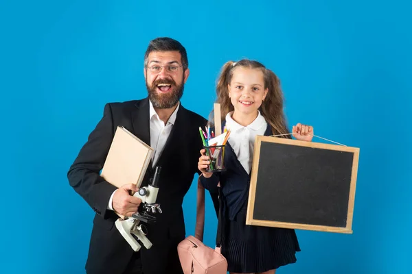 Ritratto di insegnante sorridente e allieva felice studentessa pronta a scuola. Ritratto di papà e bambine con materiale scolastico, isolate su sfondo blu. — Foto Stock