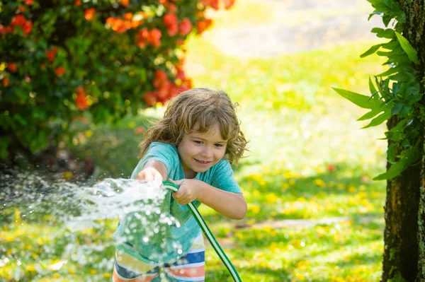 Aadorable kid boy watering the plants, from hose spray with water hose in the garden at the backyard of the house on a summer evening. — Stock Photo, Image