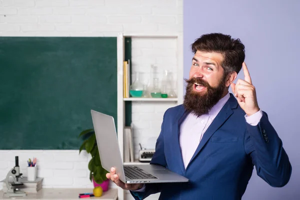 Excited teacher with pointing finger hold laptop notebook in classroom at school. Amazed teacher waiting for students, smiling confidently. Positive expression emotions funny face.