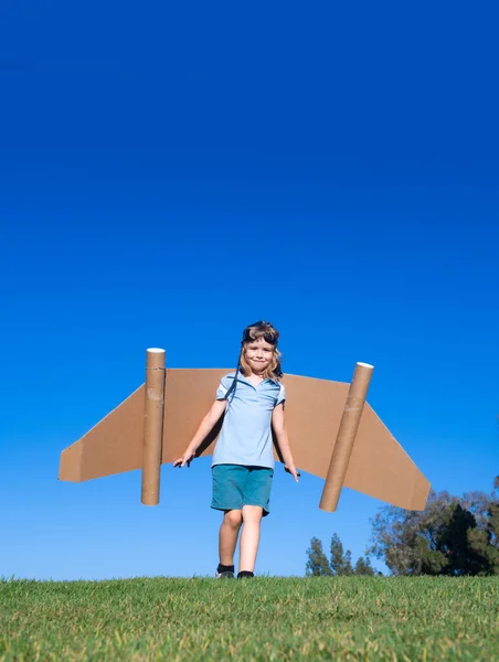 緑の芝生と青空の上で遊んでいる子供。おもちゃのジェットパック付きの子供パイロット。おもちゃの飛行機の段ボールで子供の男の子の遊び。夏の旅行と冒険。成功者、想像力、夢のコンセプト. — ストック写真