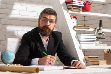 Okulda erkek öğretmenin portresi. Ciddi bir öğretmen. Masada oturuyor. Okulda ders kitabı yazıyor..