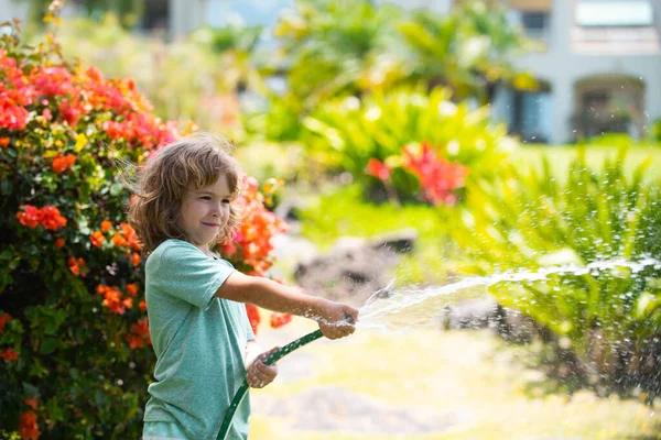 Söt liten pojke vattna blommor i trädgården på sommardagen. Barn som använder trädgårdsslang. Rolig unge vattna växter i trädgården. — Stockfoto