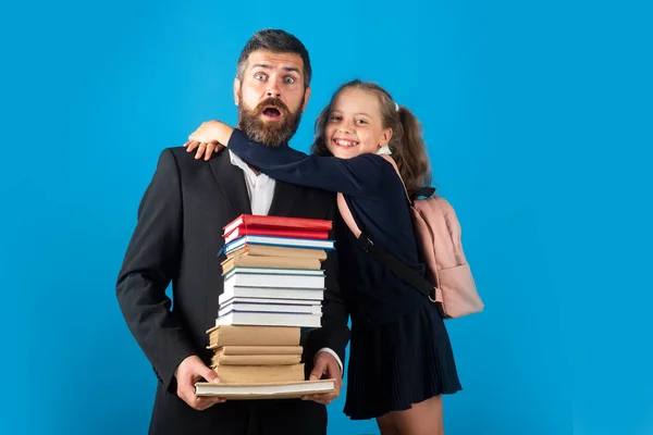 Grundschüler umarmt Lehrerin im Atelier. Lustige Vater oder Lehrer mit Schulmädchen Tochter halten großen Stapel Schulbuch Notizbücher, isoliert auf blau. — Stockfoto