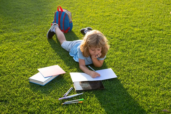 Cute boy reading book on green grass writing notes in copybook . Preparation for school and international literacy day. Back to school concept.