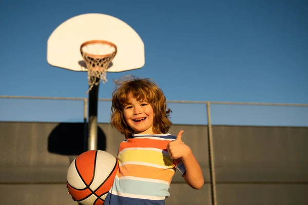 Ο χαριτωμένος χαμογελαστός παίζει μπάσκετ. Ενεργά παιδιά απολαμβάνουν υπαίθριο παιχνίδι με μπάλα καλάθι. Πορτρέτο του σπορ χαρούμενο παιδί γελώντας και διασκεδάζοντας, αντίχειρες επάνω. — Φωτογραφία Αρχείου