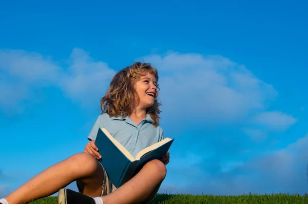 Grabben läser en bok som ligger på gräs på blå himmel bakgrund. Söt liten barn i casual kläder läser en bok och ler när du ligger på gräs i parken. — Stockfoto