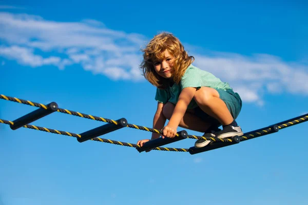 Rapaz bonito sobe a escada no parque infantil. Criança sobe a escada contra o céu azul. Bonito menino bonito sorridente em um playground. Actividades infantis. — Fotografia de Stock