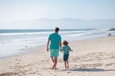 Baba ve çocuk el ele tutuşup birlikte yürüyorlar. Baba oğul yaz plajında yürüyorlar..
