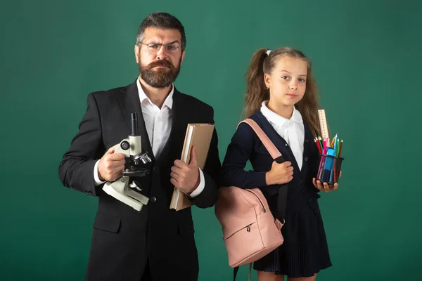Portrait of serious teacher and pupil school girl ready to school. Portrait of dad and little girls with school supplies, isolated on green blackboard background. — Foto Stock