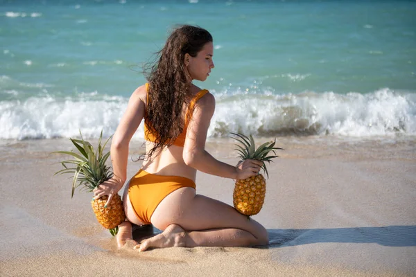 Giocoso donna magra in bikini sexy si erge indietro cammina spiaggia di mare con ananas fresco in mano. Calda spiaggia estiva, costumi da bagno per donne. — Foto Stock