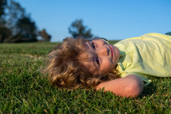 Porträtt av en glad liten pojke som ligger på gräset i parken. Utomhus porträtt av söt liten pojke på färskt grönt gräs. Sommar kul, lycklig barndom. — Stockfoto