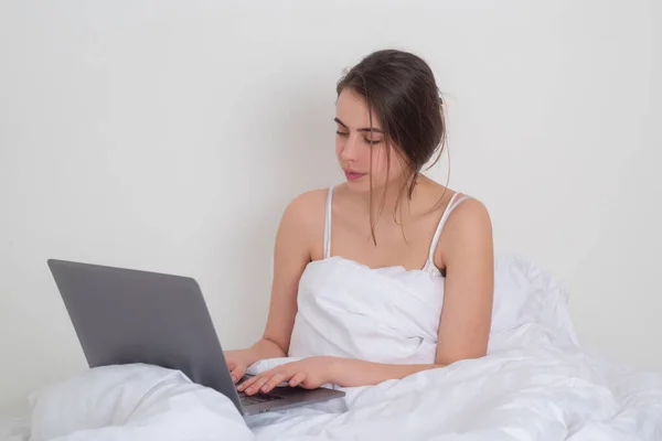 Bella donna con computer portatile a letto. Modello bruna con laptop in pigiama. Una donna lavora al computer nella sua camera da letto a casa. — Foto Stock